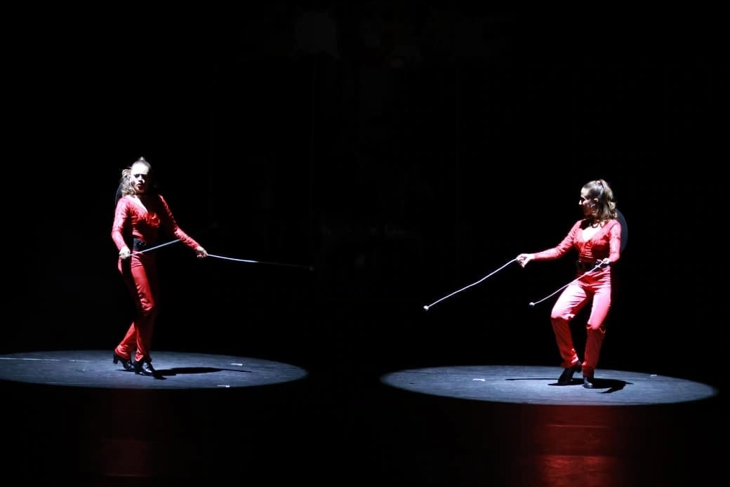 Two dancers in red costumes performing with boleadoras under separate spotlights on a dark stage